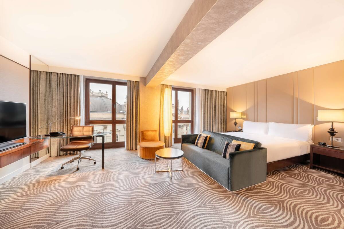 A hotel room with a bed, a sofa, a desk, a chair, and a TV.