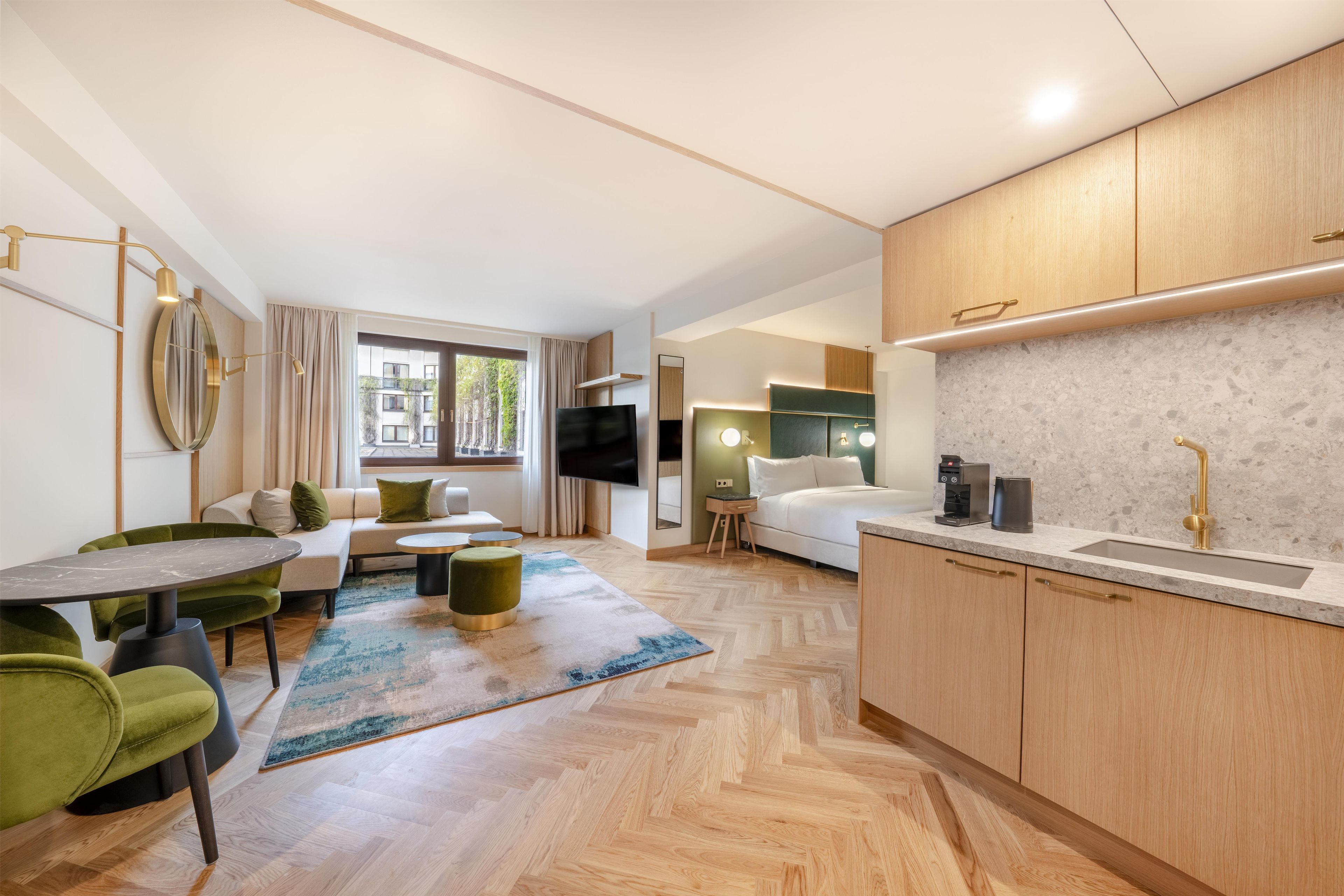 A modern hotel room with a sofa, dining table, kitchen area, and a large TV.
