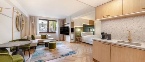 A modern hotel room with a sofa, dining table, kitchen area, and a large TV.