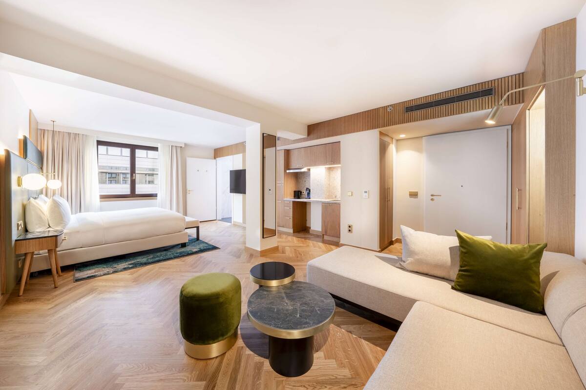 A modern hotel room with a large bed, a sofa, a small round table, and a green ottoman. There is a kitchen area with cabinets and a countertop visible through an open door.