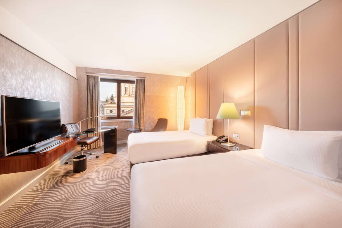 A hotel room with two beds, a TV, a desk with a chair, and a lamp.