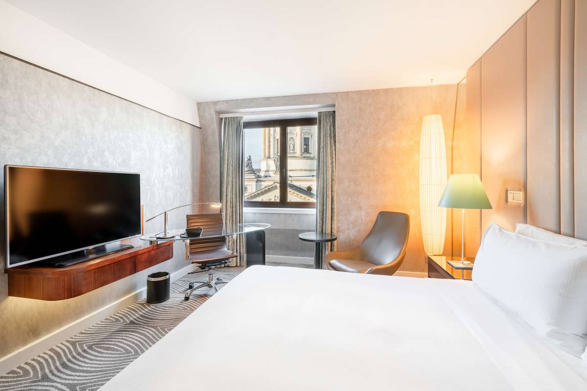 A hotel room with a large bed, a flat-screen TV, a small table, a chair, and a view of a cityscape through a large window.