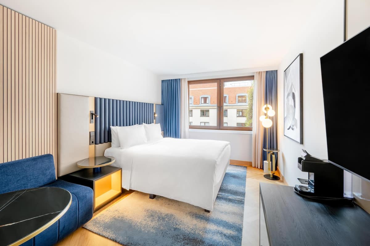 A modern hotel room with a bed, a sofa, a desk, a TV, and a window with curtains.