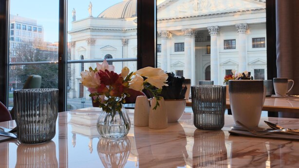A table with a vase of flowers, salt and pepper shakers, and a bowl. A large window reveals a classical building with columns and a dome.