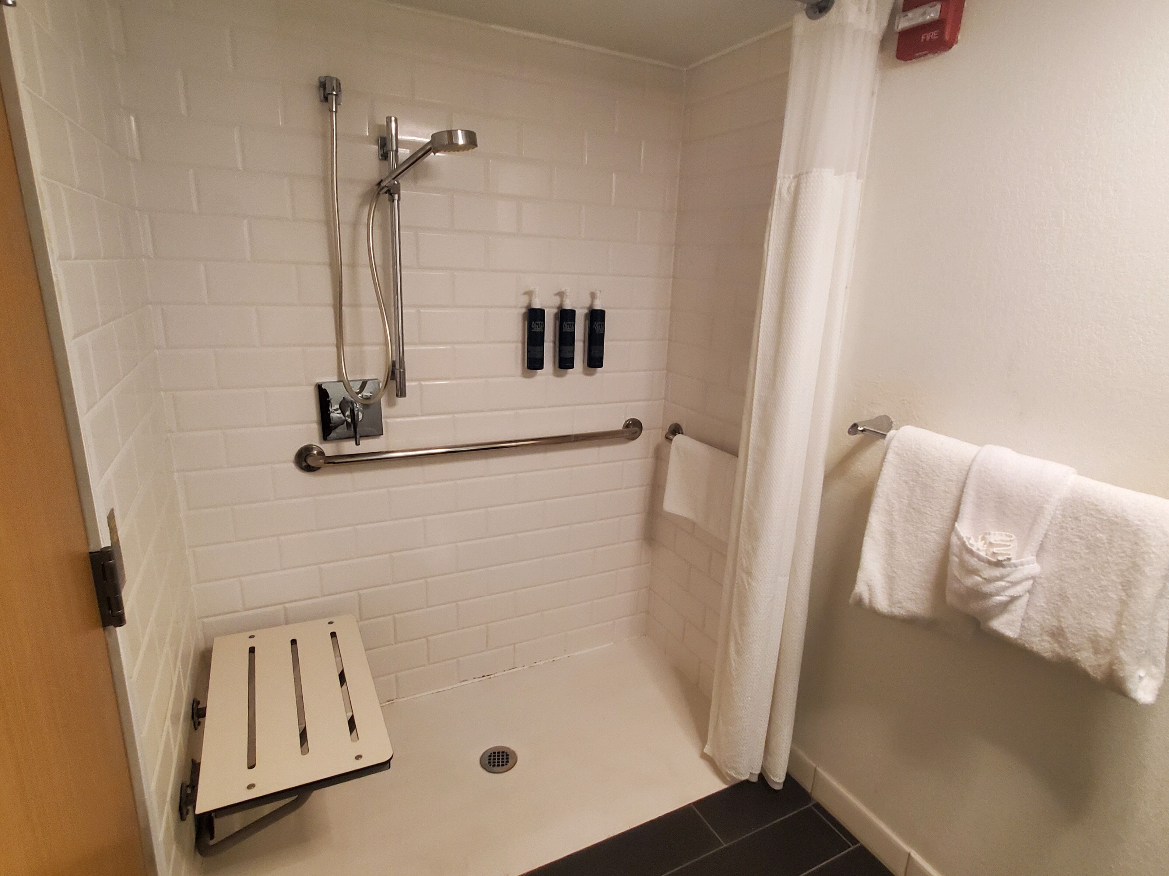 Un petit espace de douche fermé, équipé d’une barre d’appui, d’un porte-serviettes et d’un mur recouvert de carreaux blancs.