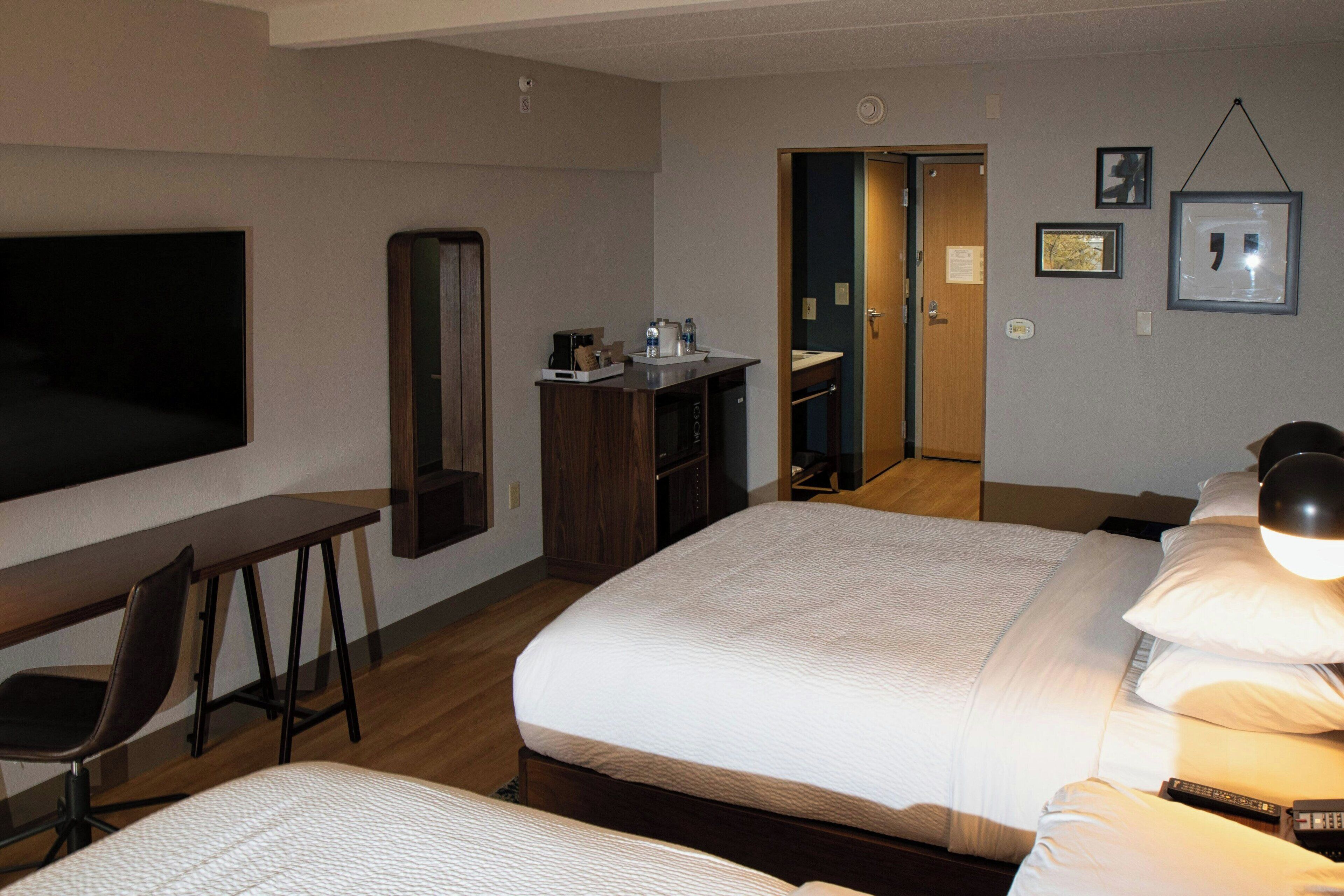 A hotel room with two beds, a desk, a chair, a TV, a mirror, and a door leading to a bathroom.