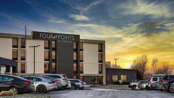 A modern building with a sign reading "FOUR POINTS BY SHERATON" and a parking lot with several cars.