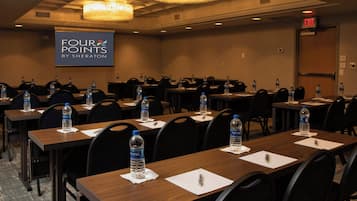 A conference room with multiple tables, chairs, and water bottles. A projector screen displays the text "FOUR POINTS BY SHERATON".