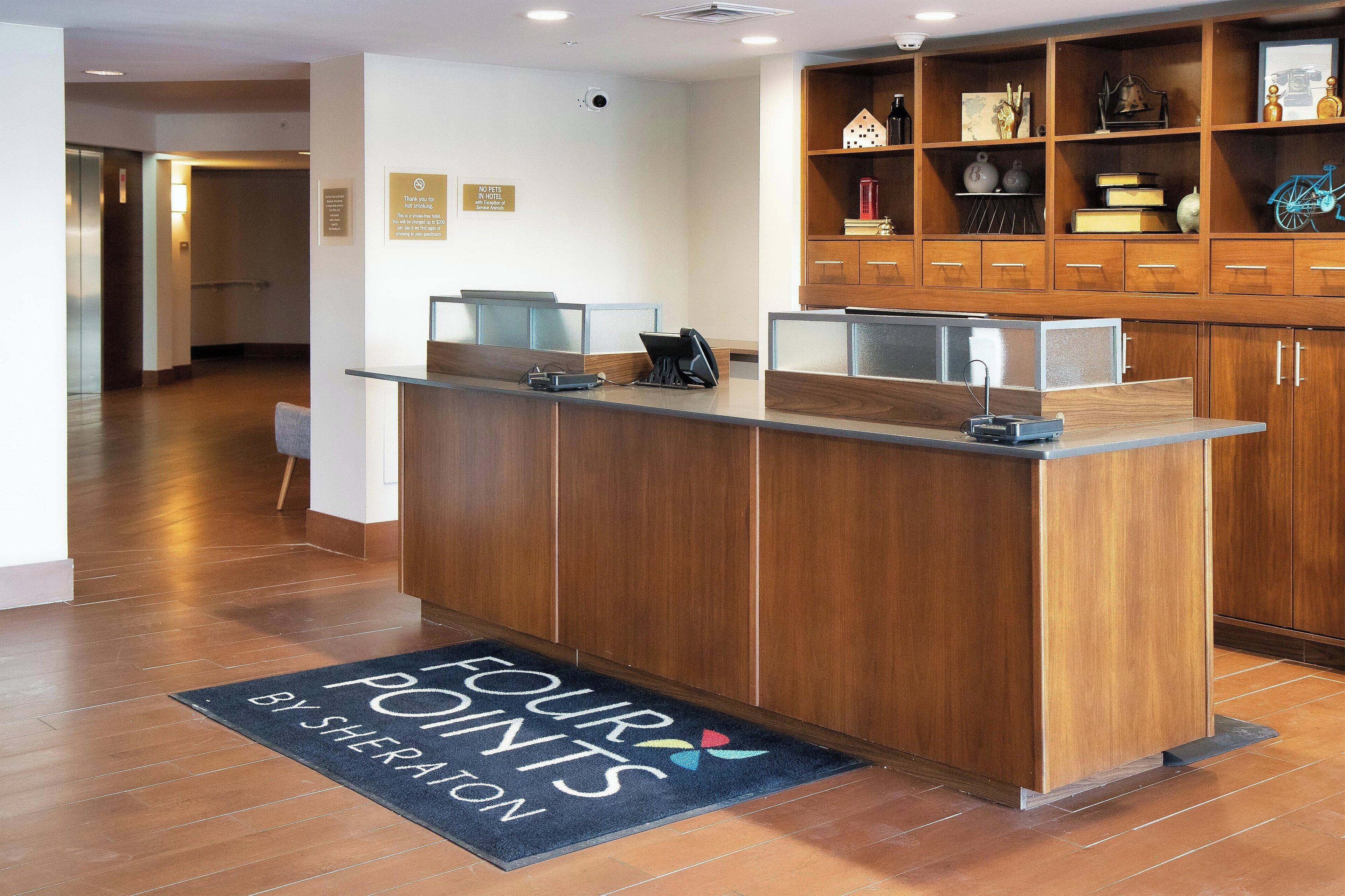 A reception desk with a wooden finish, a glass partition, and a black doormat with "FOUR POINTS BY SHERATON" text.