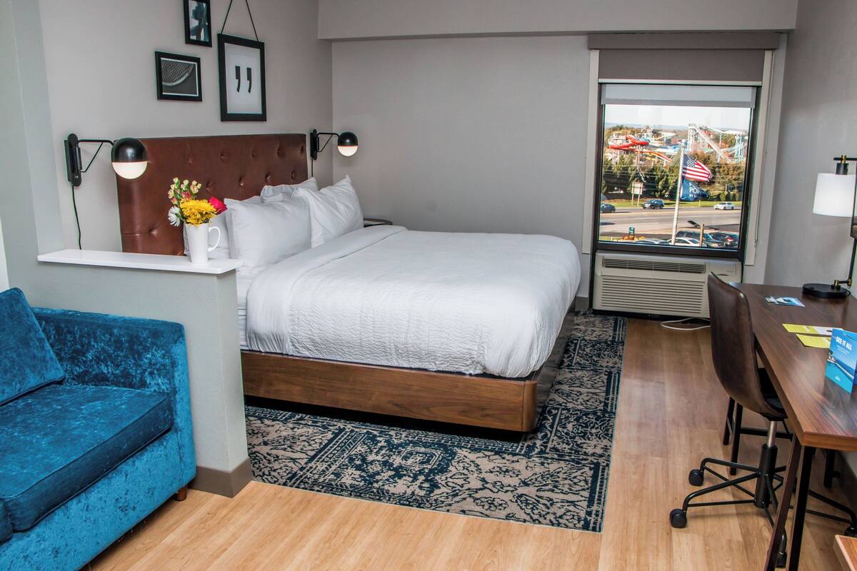 Hotelkamer met een bed, een blauwe bank, een bureau met stoel, een raam met een Amerikaanse vlag en een wandplank met een bloevas.