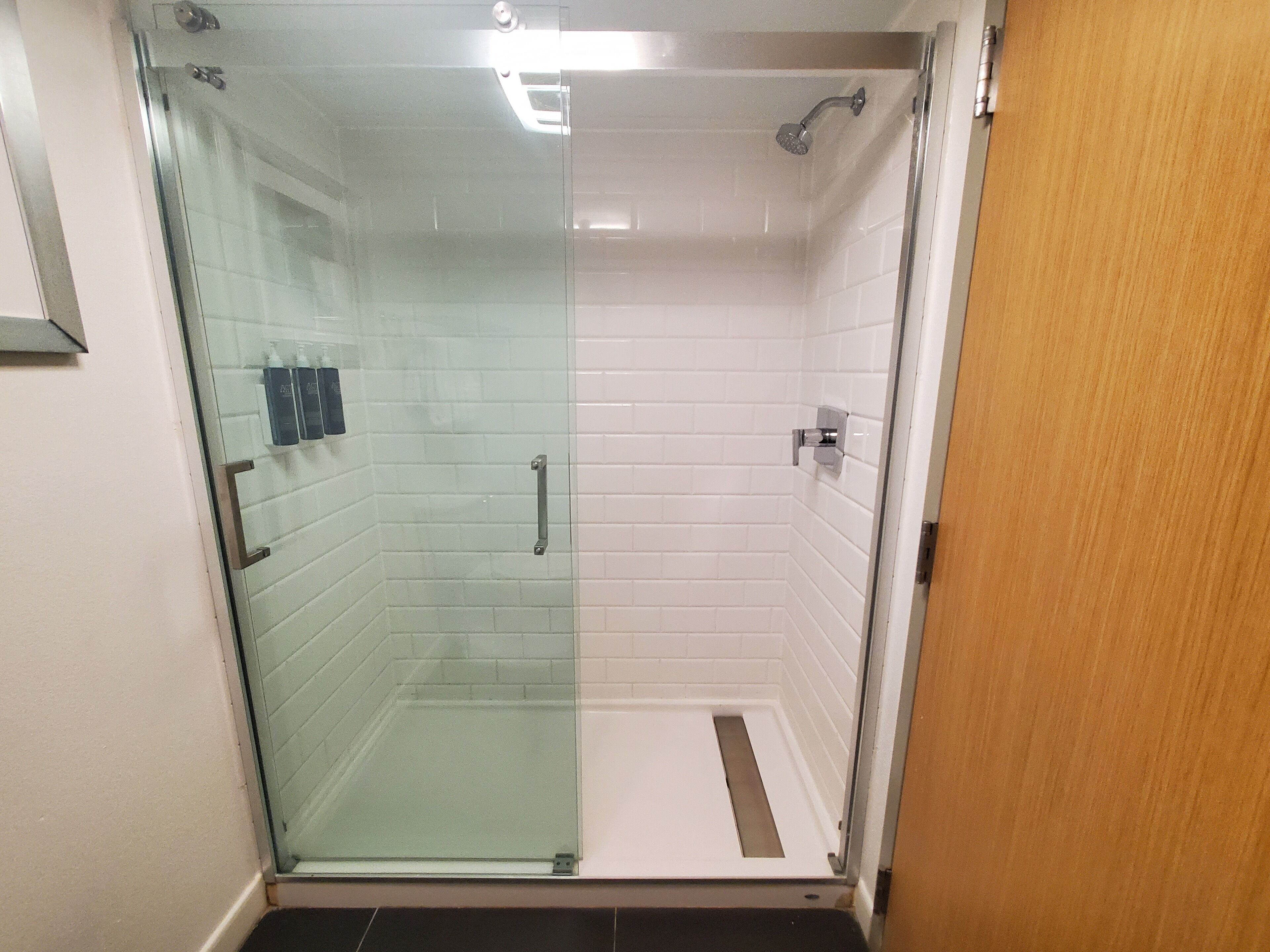 A glass-enclosed shower stall with white tiled walls and a wooden door.