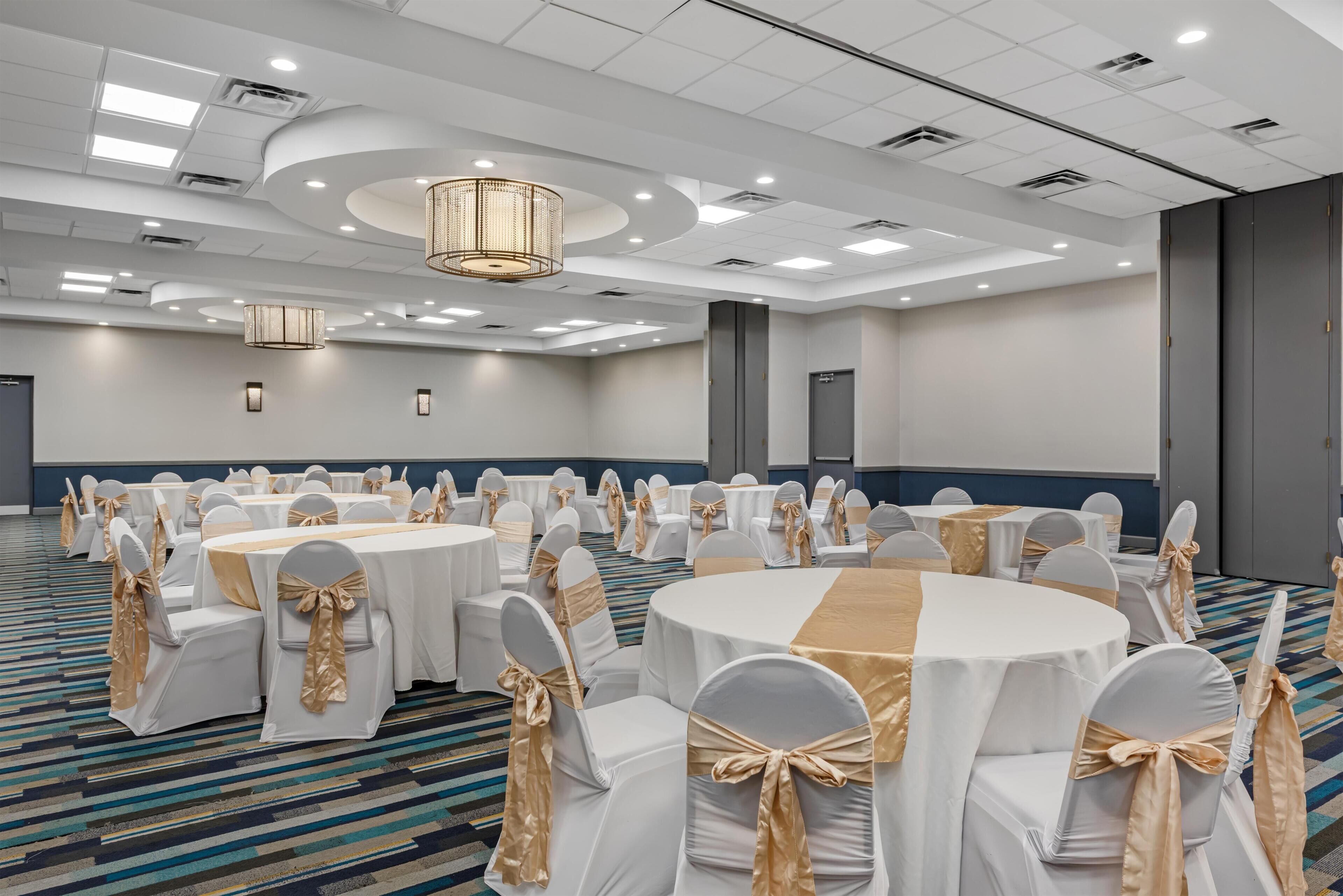 Un salón de conferencias con mesas redondas, sillas blancas con cintas doradas e iluminación empotrada en el techo.