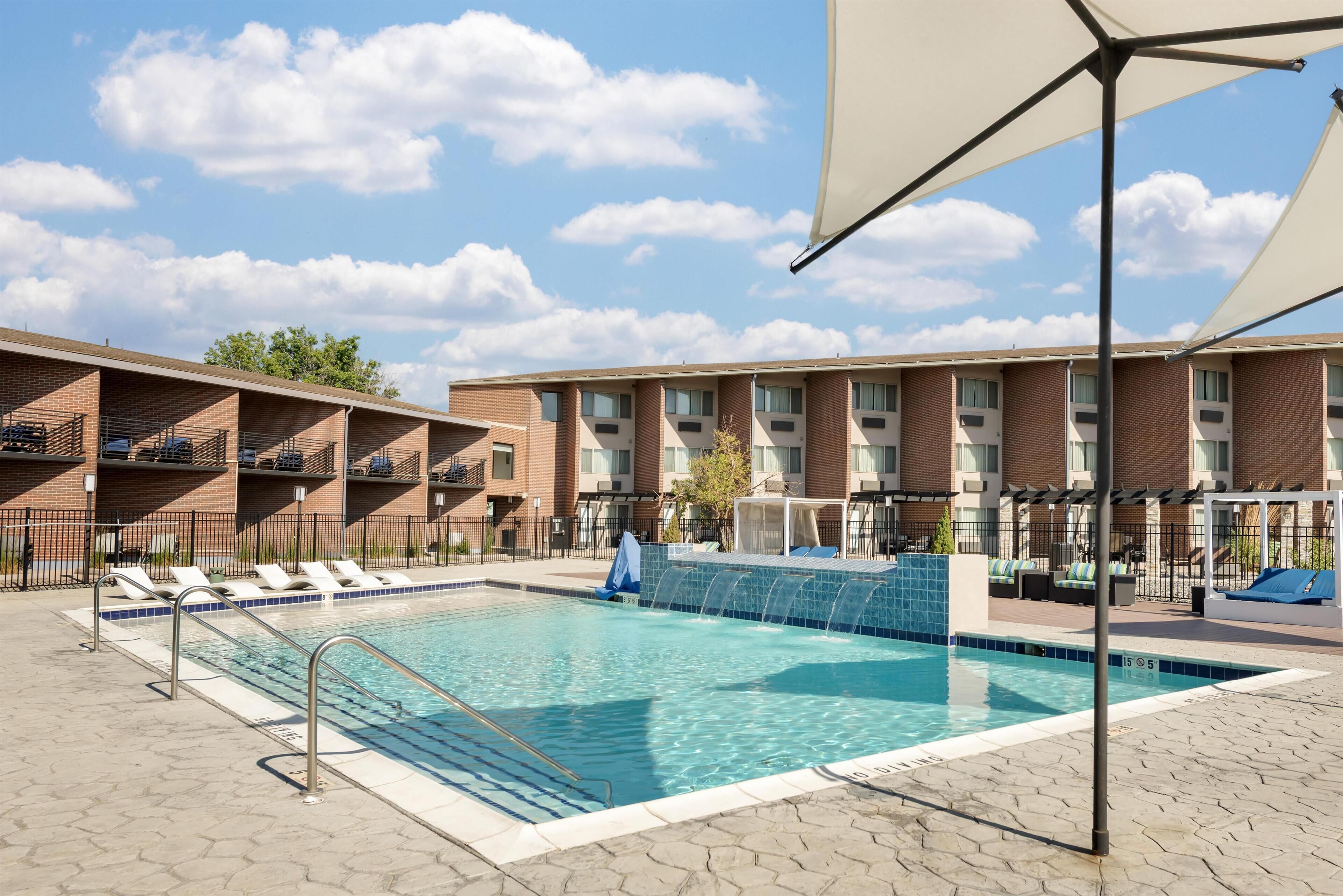 Una piscina con sillones de descanso, una zona cubierta y un edificio de ladrillo con varios balcones.