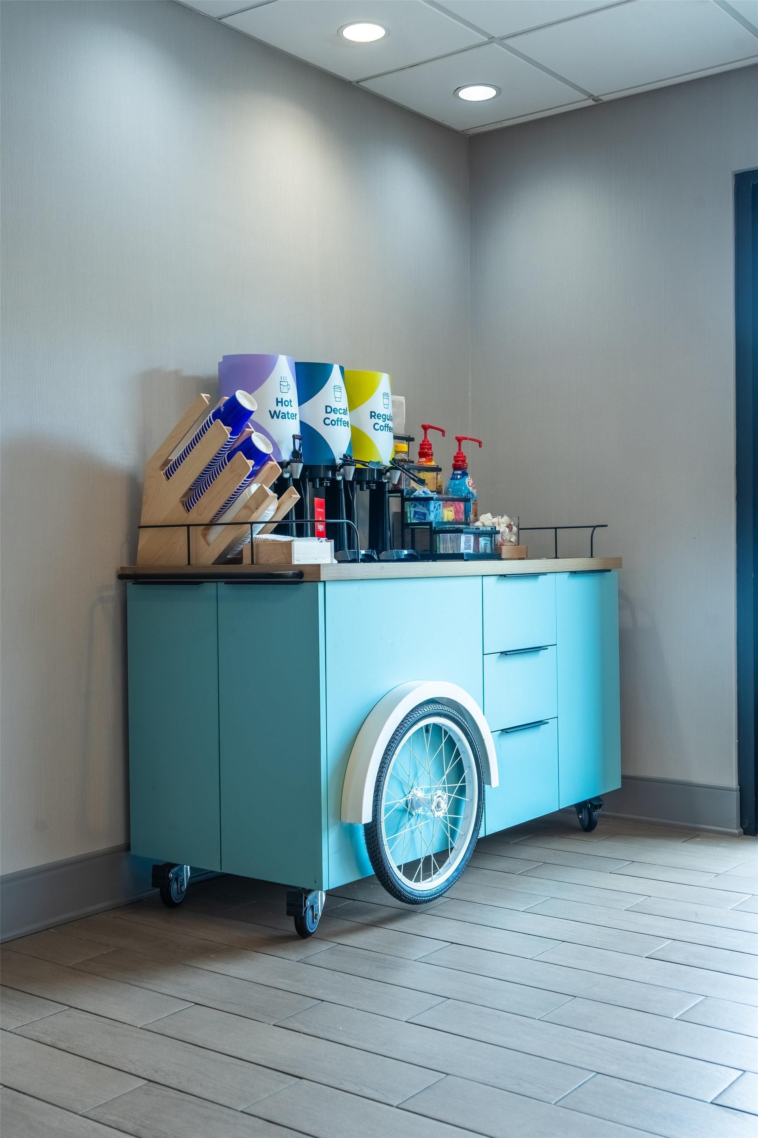 A teal mobile cart with a wheelchair wheel, containing various items like coffee machines and bottles.