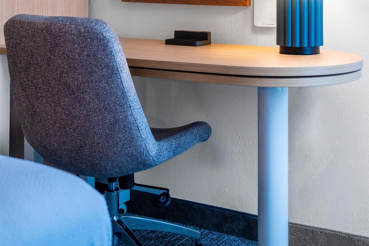 A hotel room with a desk, a chair, a lamp, a framed picture on the wall, and a television.