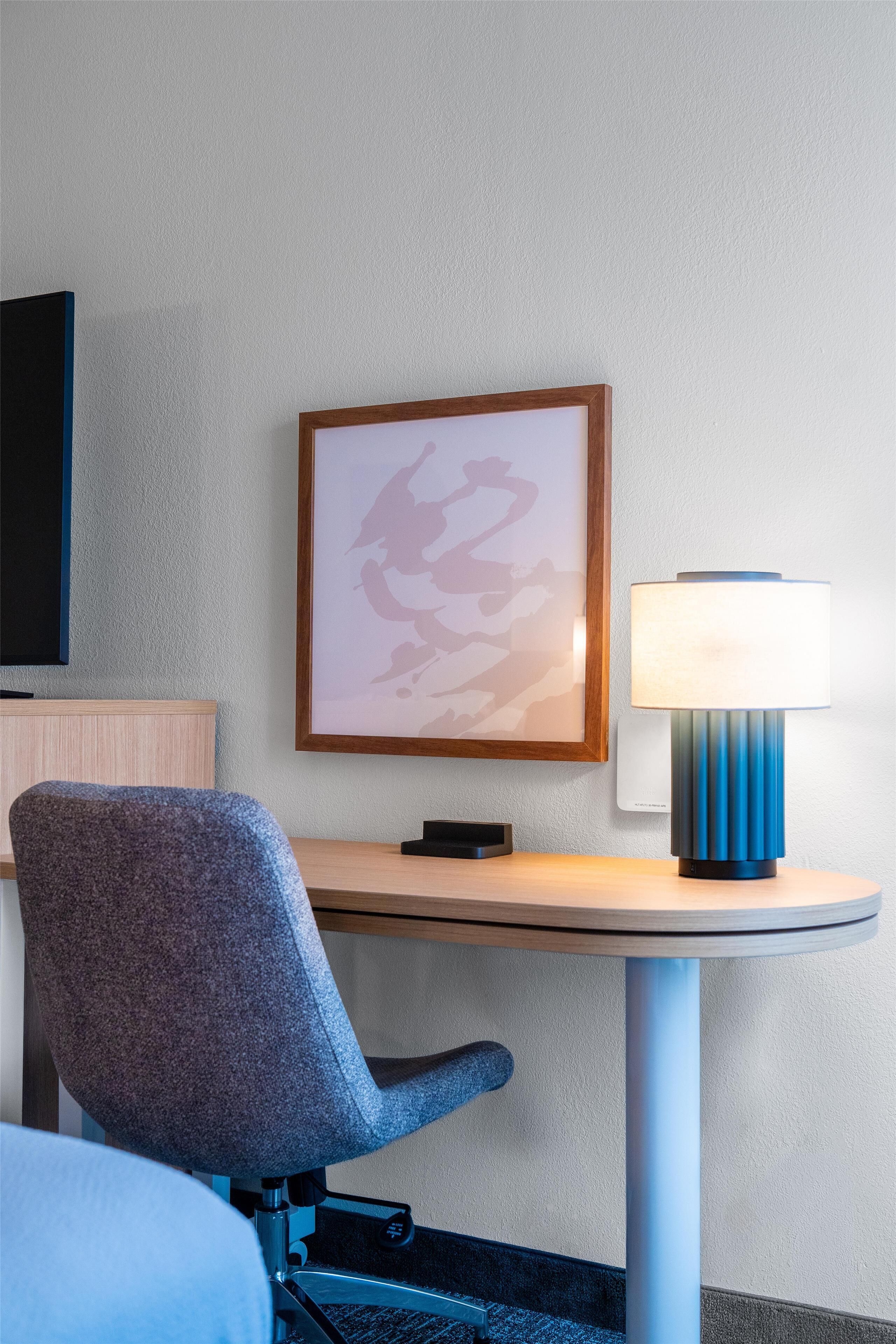 Una camera d'albergo con una scrivania, una sedia, una lampada, un quadro appeso al muro e un televisore.