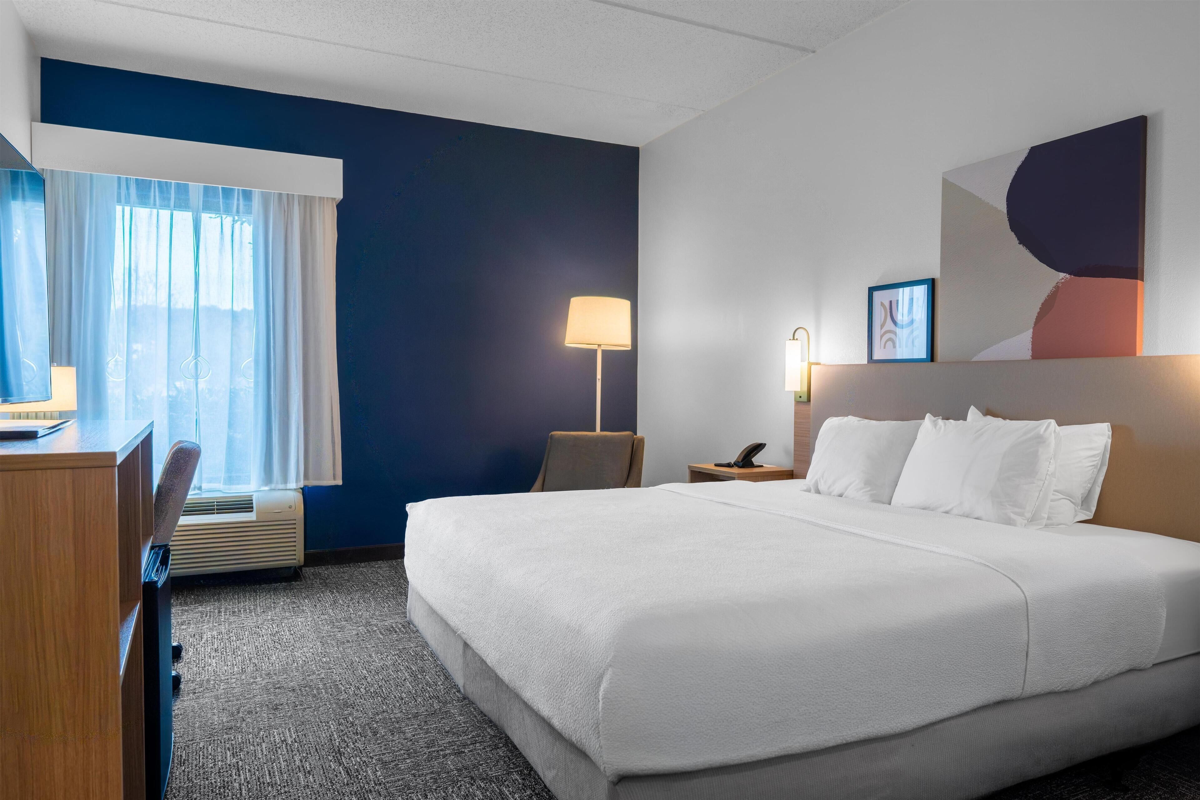Une chambre d’hôtel avec un lit, un bureau, une chaise, une lampe et une œuvre d’art au mur.