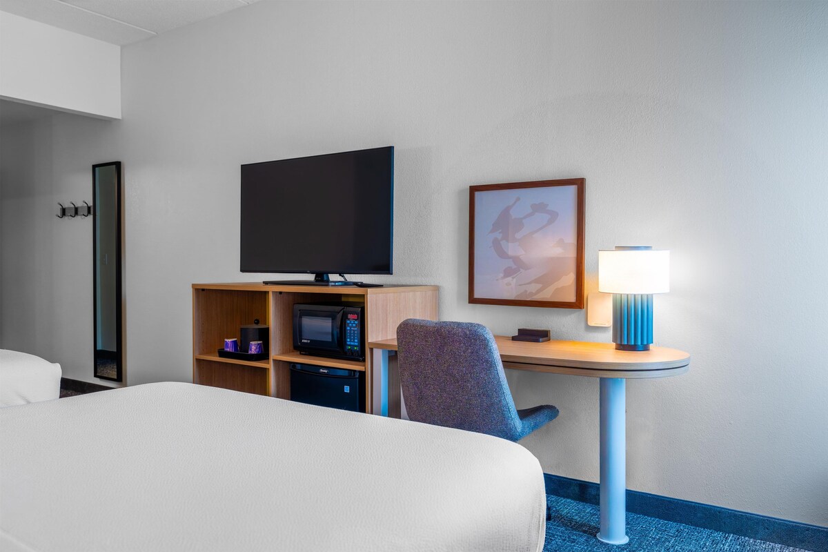 A hotel room with a bed, a desk with a television, a microwave, a chair, a lamp, and a framed picture on the wall.