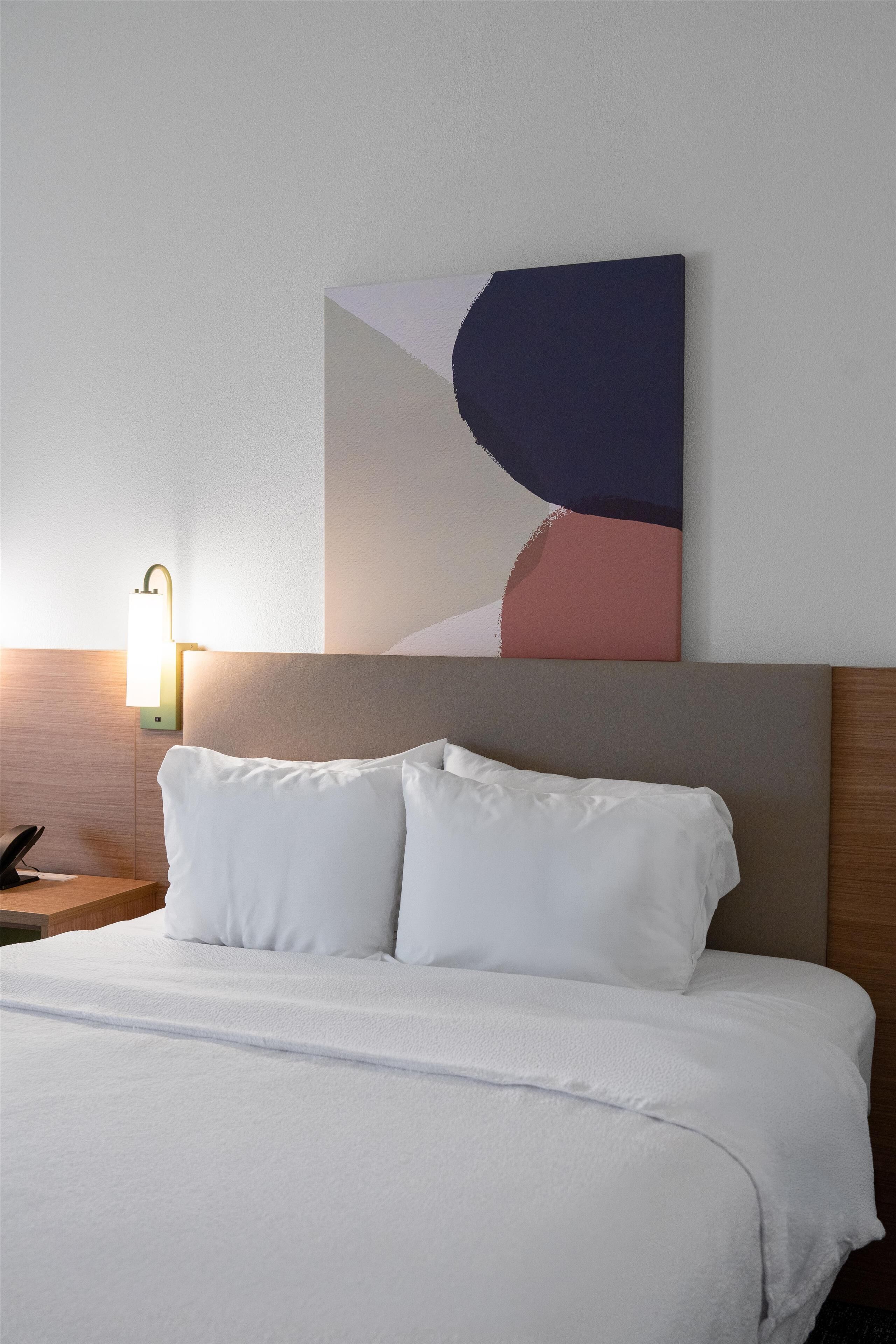 Ein ordentlich bezogenes Bett mit weißen Leinen, ein an der Wand befestigtes abstraktes Gemälde und eine Nachttischlampe.