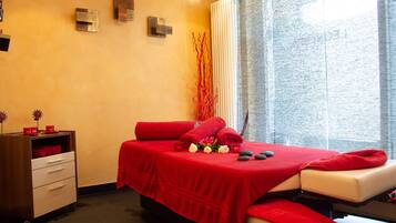 A spa room with a red bed, a massage table, a wooden cabinet, and a window with curtains.