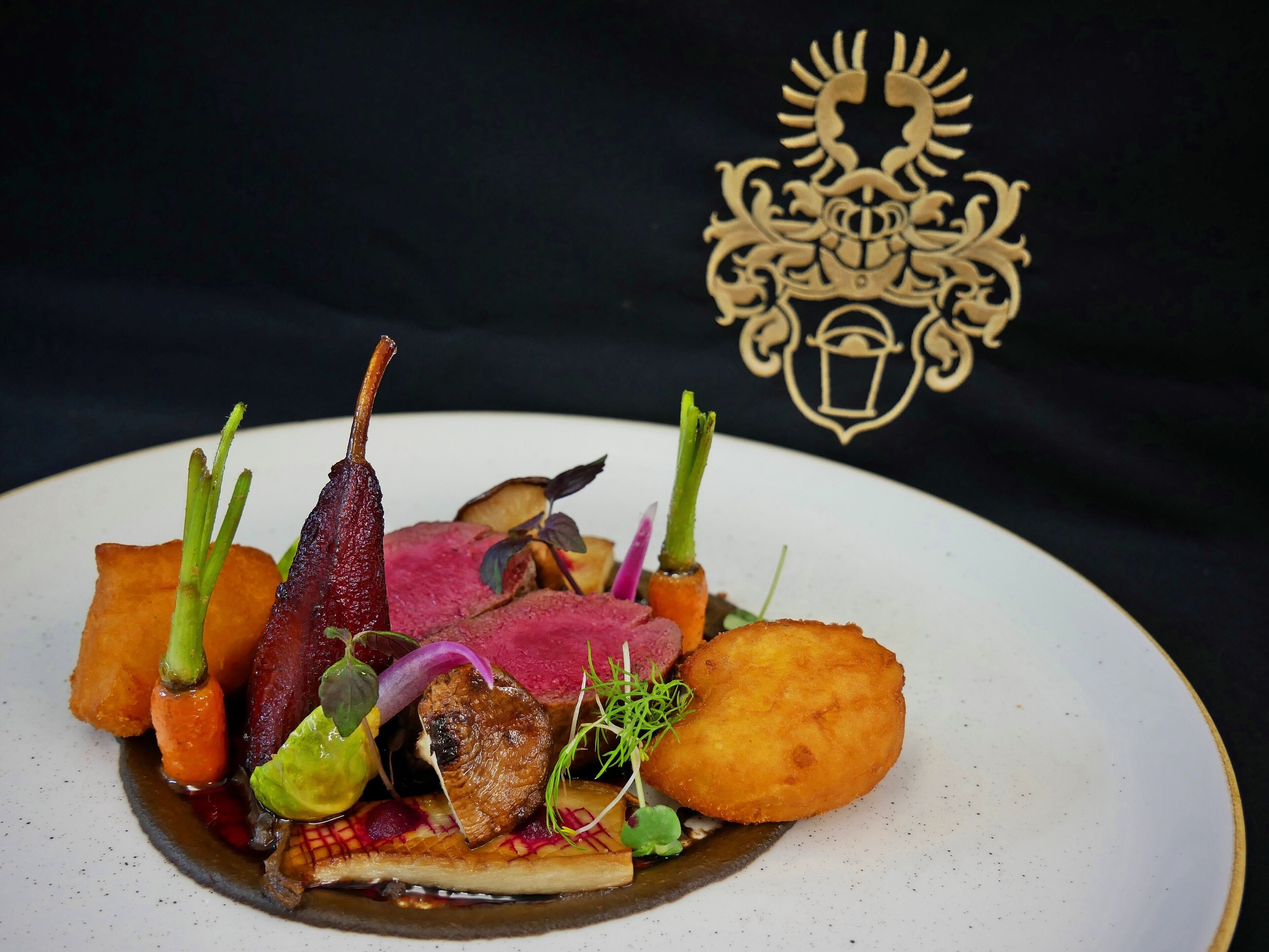 Prato gourmet com diversos elementos, incluindo carne, legumes e guarnições, apresentado em um prato branco sobre fundo preto.