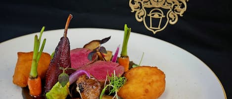 Prato gourmet com diversos elementos, incluindo carne, legumes e guarnições, apresentado em um prato branco sobre fundo preto.