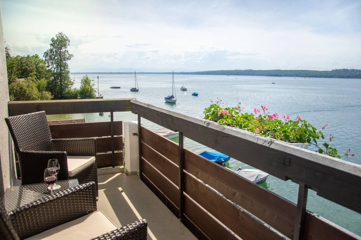 A balcony with wicker furniture, a glass of red wine, and a view of boats on a lake.