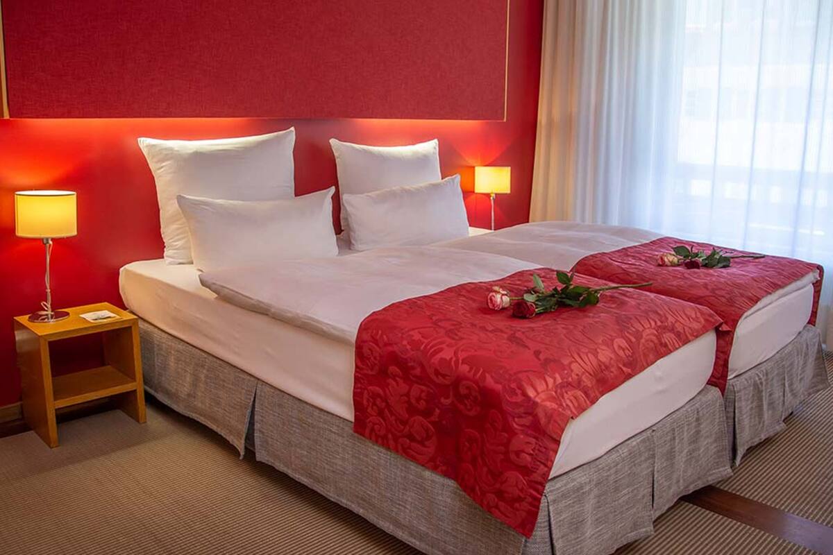 A hotel room with a large bed, two bedside lamps, a red headboard, and a bouquet of flowers on the bed.