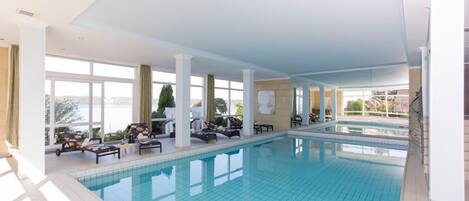 An indoor swimming pool with tiled flooring, sunlit areas with large windows, and lounge chairs.