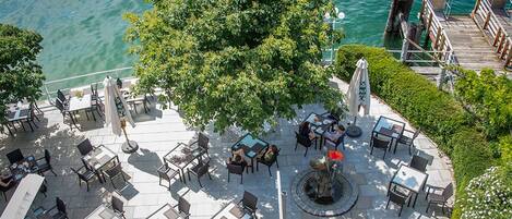 Un patio al aire libre con mesas y sillas, un árbol grande y vista al agua.
