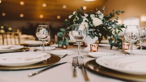 A dining table set with white plates, gold cutlery, and clear wine glasses, surrounded by greenery and copper candle holders. - Belamar Hotel Manhattan Beach, Tapestry Collection by Hilton (Manhattan Beach)