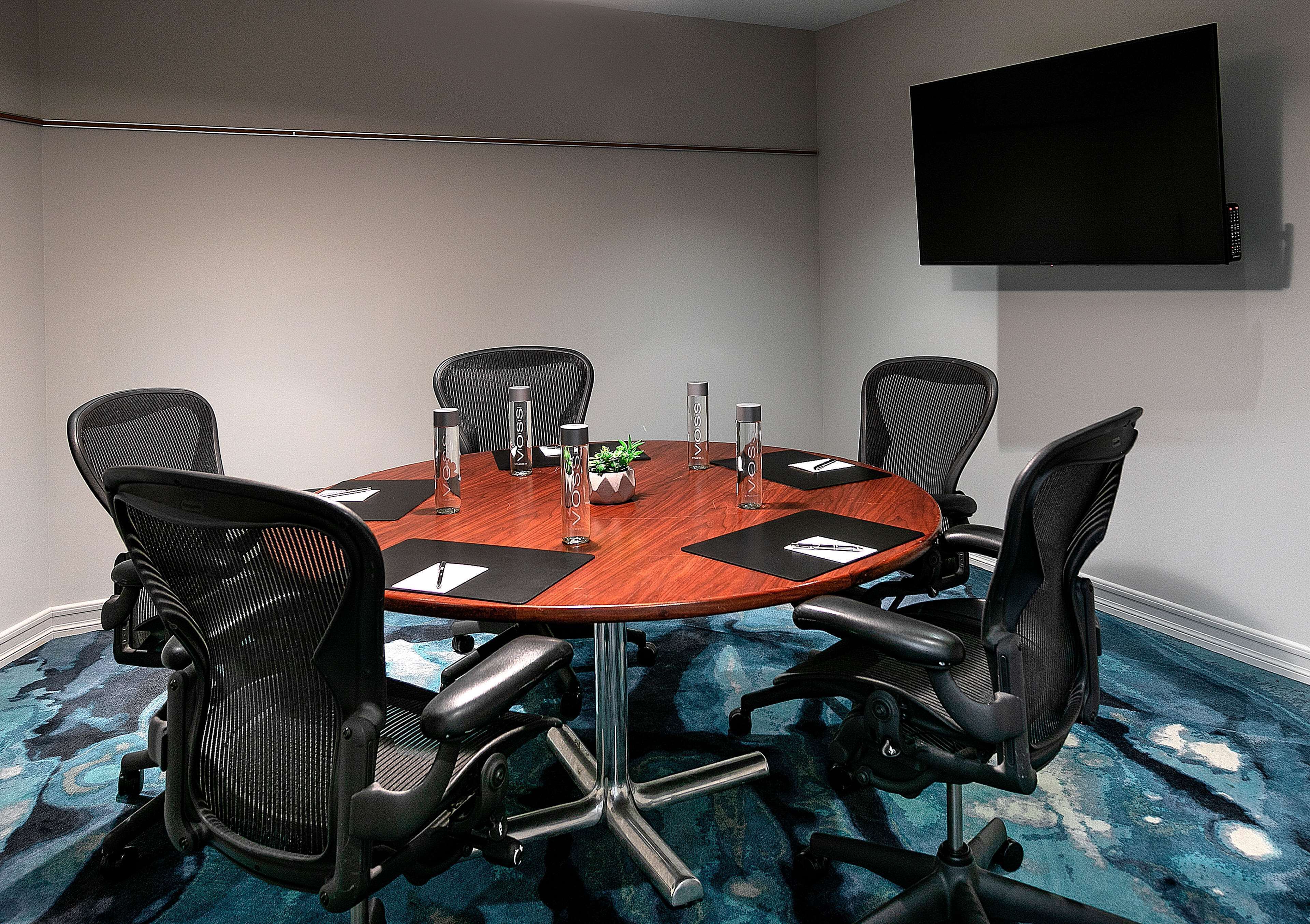 Uma sala de conferências com uma mesa redonda, seis cadeiras de escritório pretas, uma TV de tela plana na parede e um carpete azul com padrão discreto.