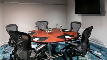 Uma sala de conferências com uma mesa redonda, seis cadeiras de escritório pretas, uma TV de tela plana na parede e um carpete azul com padrão discreto.
