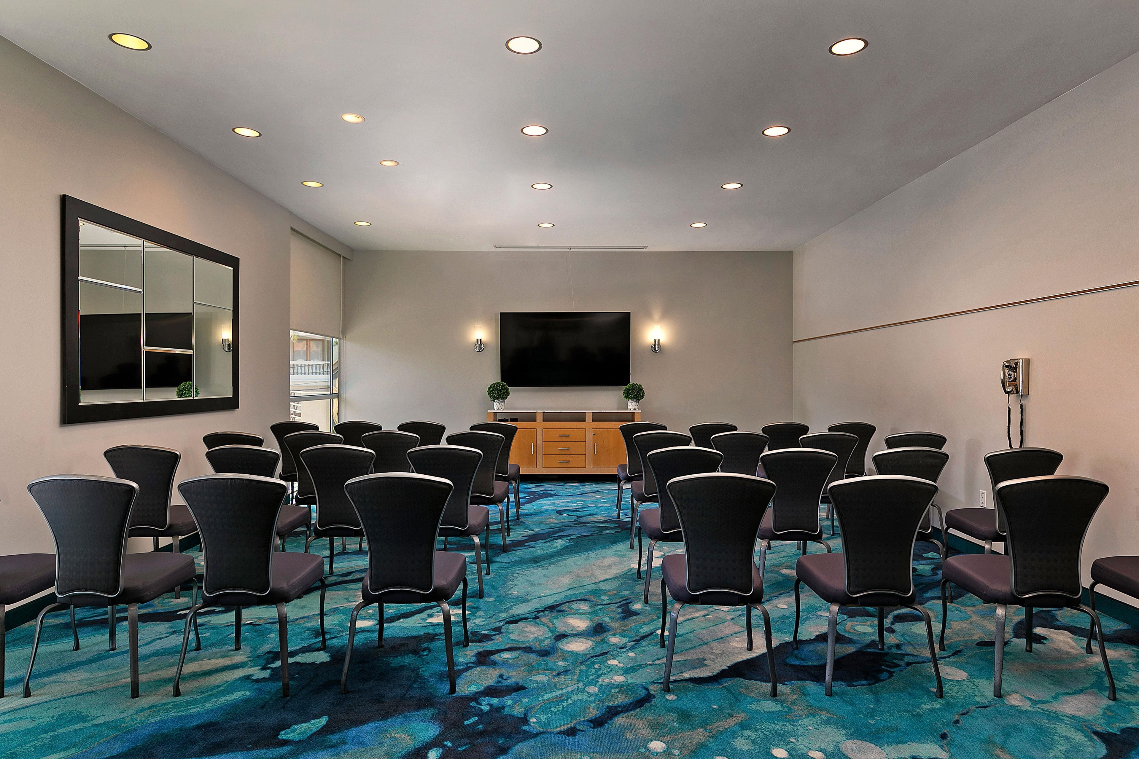 A conference room with rows of chairs facing a large screen, a wooden cabinet, and a mirror on the wall.