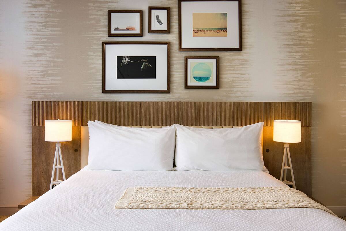Een netjes opgemaakt bed met witte lakens, twee bedlampjes, een houten hoofdbord en ingelijste foto's aan de muur.