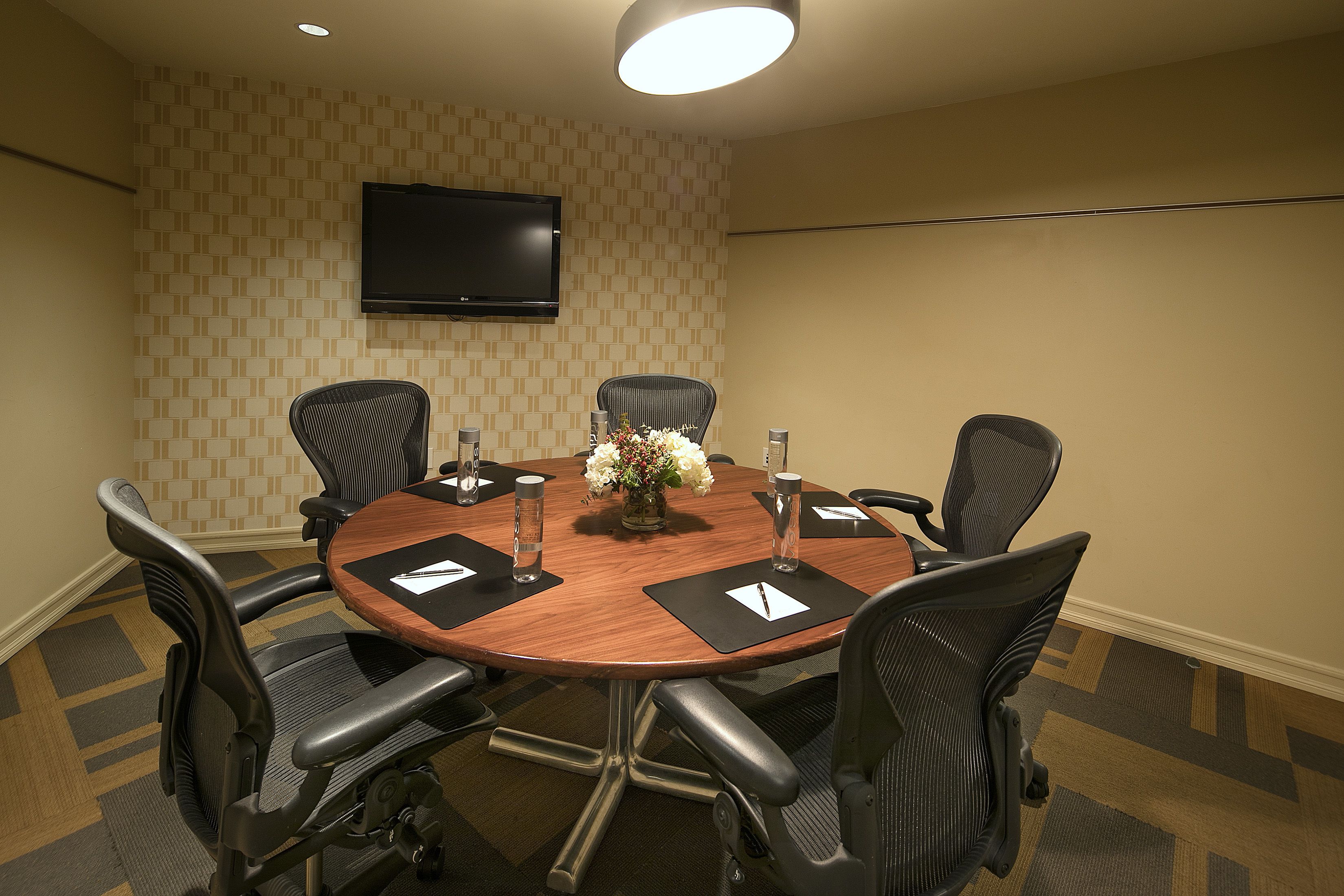 A conference room with a round table, six chairs, a flat-screen TV on the wall, and a vase with flowers on the table.