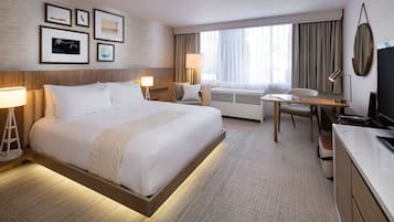 A hotel room with a large bed, bedside tables, a desk, a chair, a TV, and a large window with curtains.