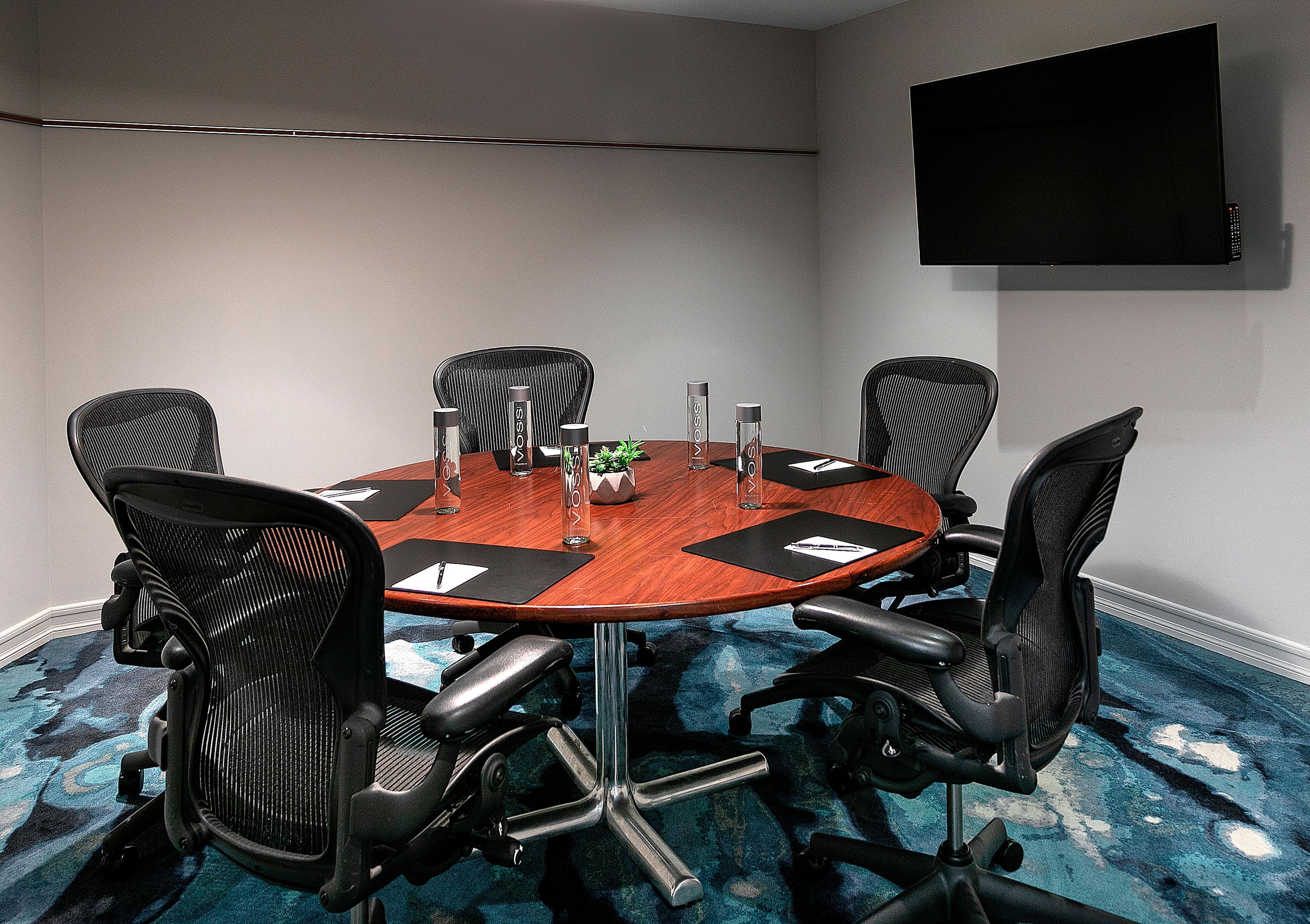 A conference room with a round table, six chairs, a flat-screen TV on the wall, and a carpeted floor.