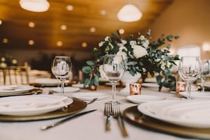 Mesa de comedor dispuesta con platos blancos, cubiertos y copas de vino, con un centro de mesa floral.