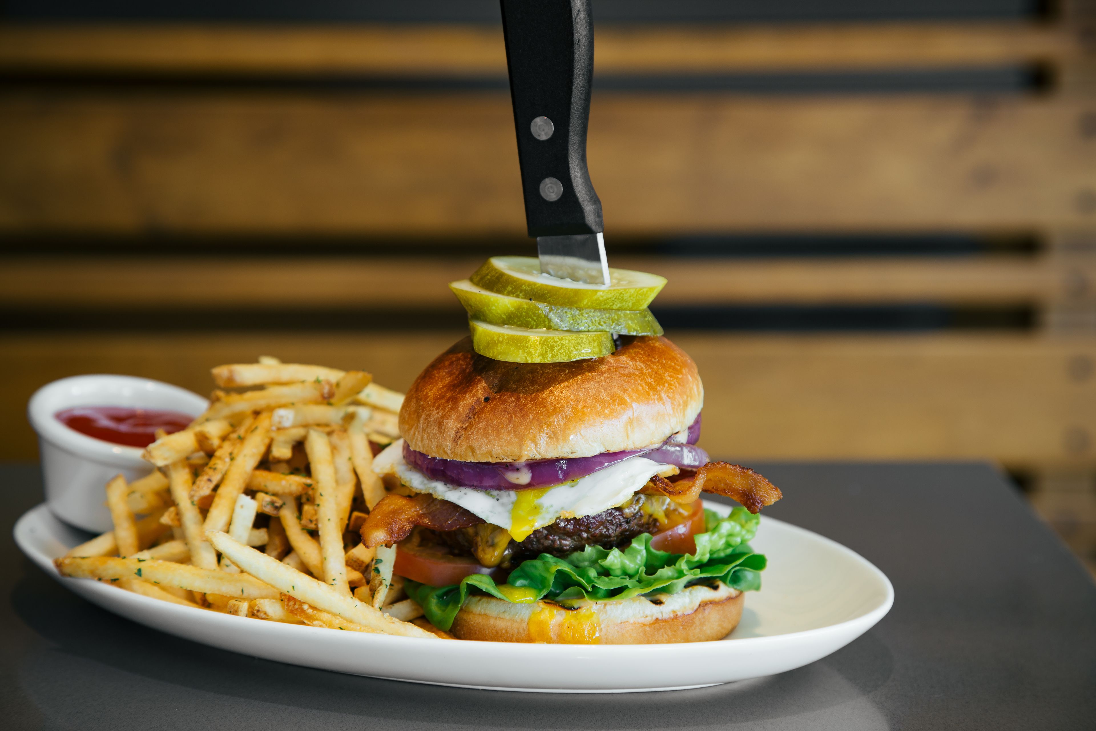 A plate with a burger, fries, and a knife cutting through a pickle.