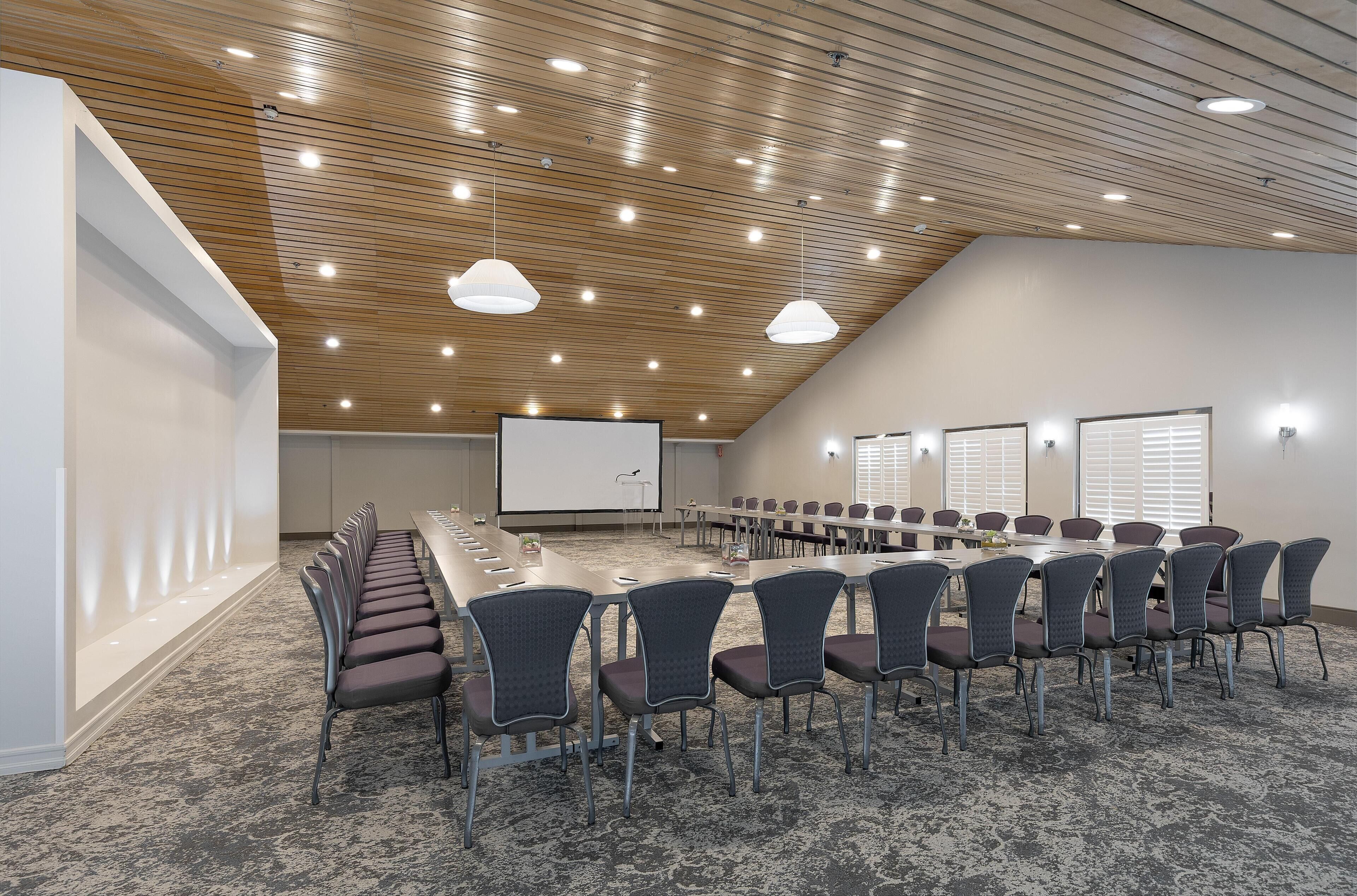 A conference room with a long table, chairs, a projector screen, and a sloped ceiling with recessed lighting.