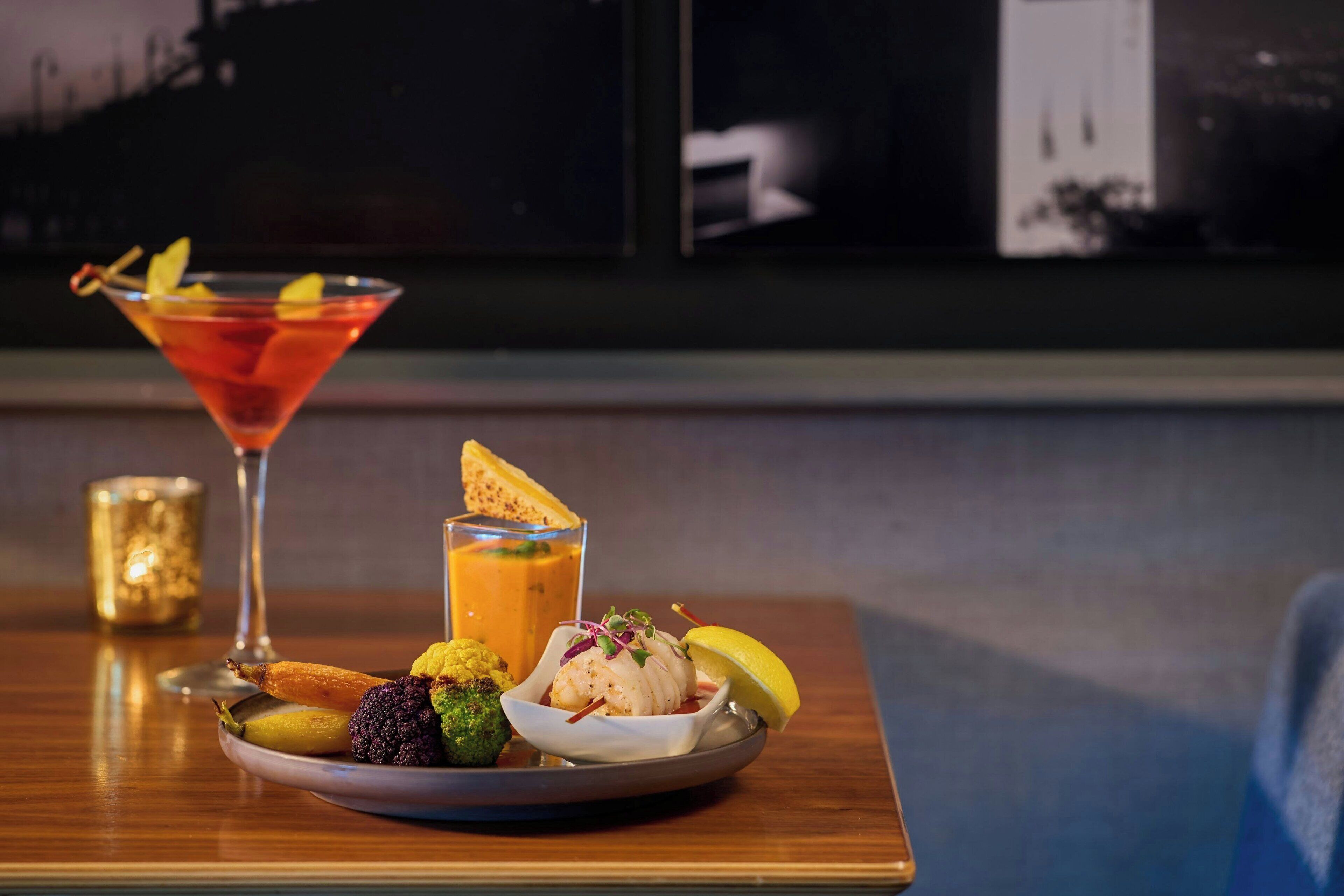 Een eettafel met een martini-glas, een klein glas sinaasappelsap en een bord met diverse hapjes.