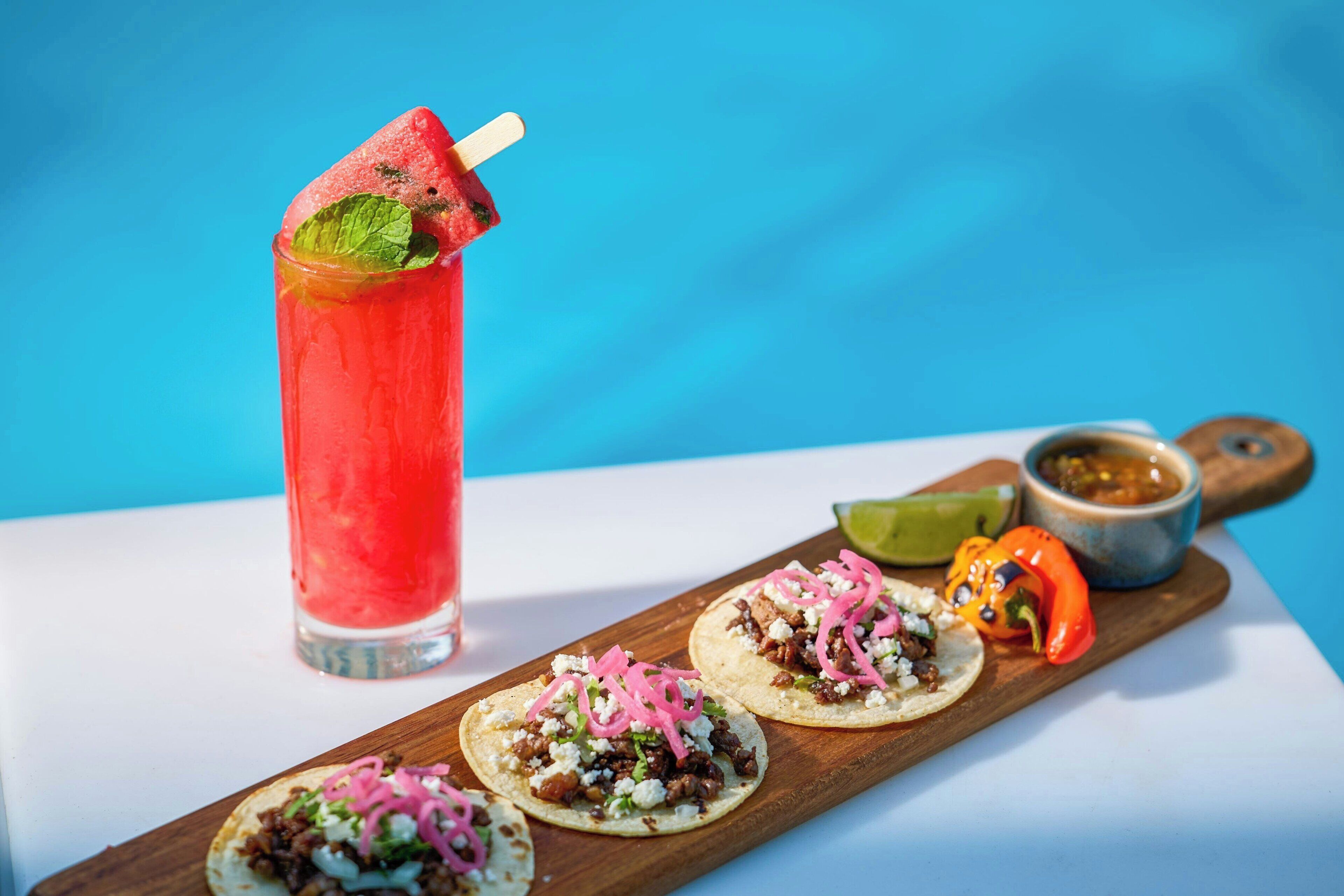 Une planche en bois sur laquelle sont disposés trois tacos et un grand verre d’une boisson rouge, garnie d’une feuille de menthe.