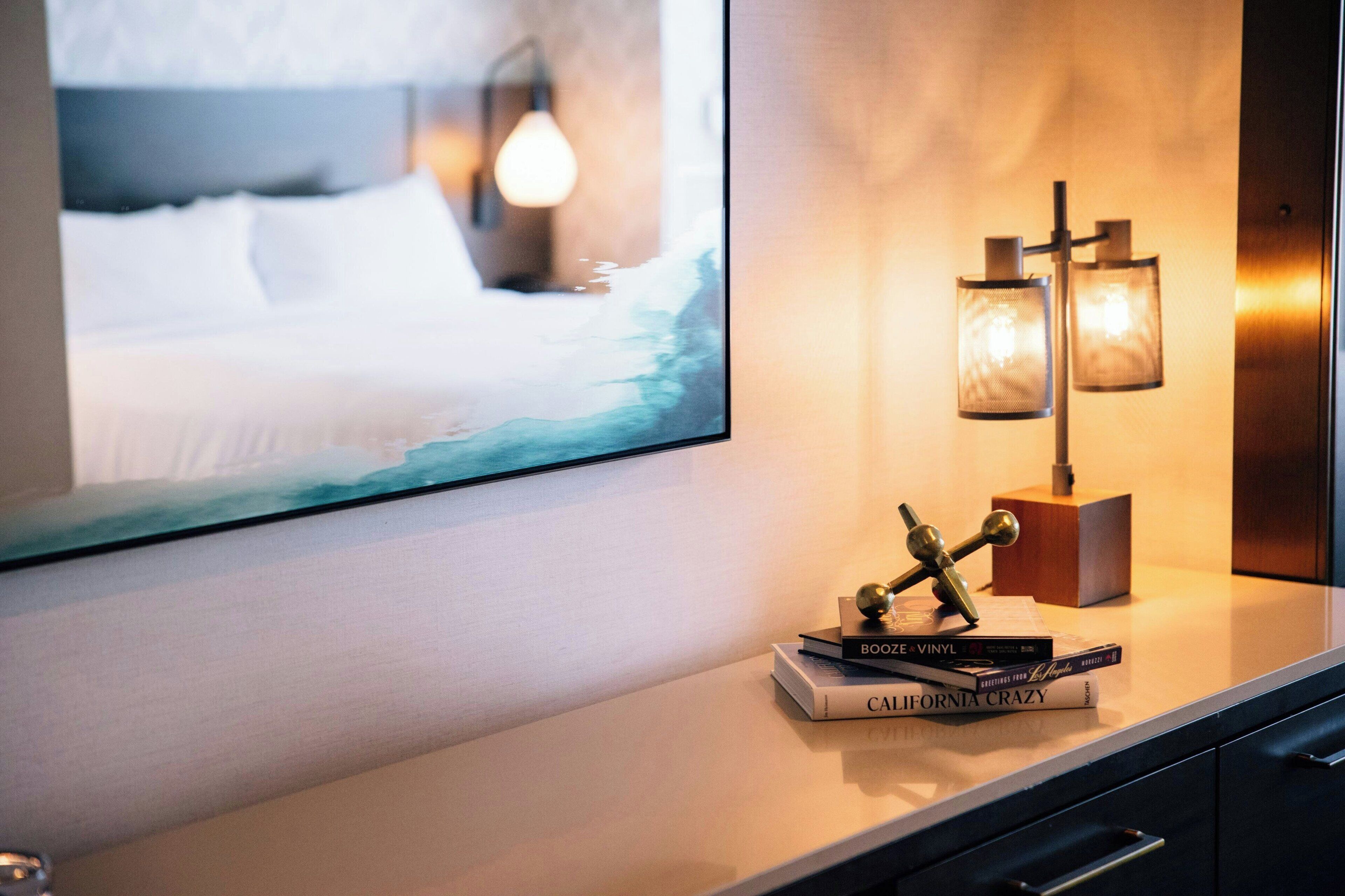 A hotel room with a bed, a wall-mounted TV, a bedside table with books and a decorative item, and a wall-mounted lamp.