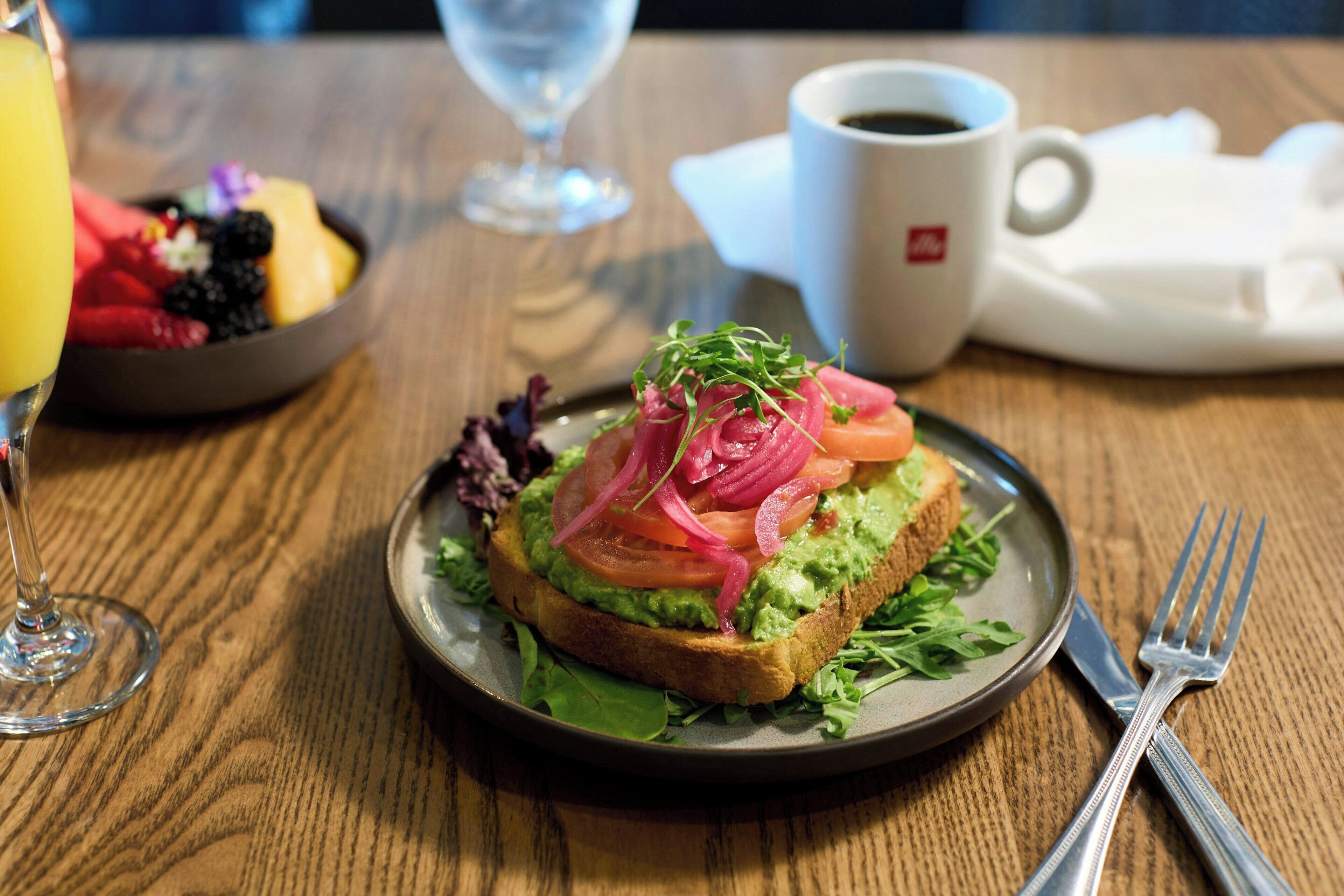Un plato de desayuno con tostadas, aguacate, tomate, vegetales de hoja, un vaso de jugo de naranja, una taza de café y un tazón de fruta sobre una mesa de madera.