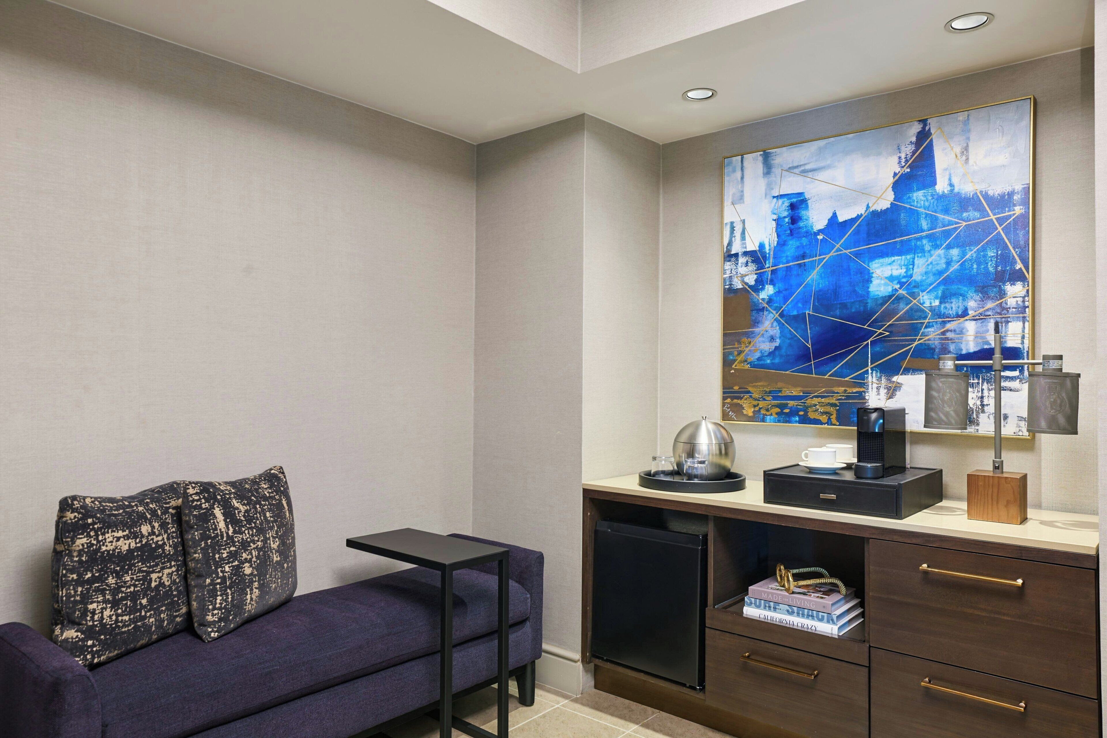 A modern hotel room with a dark blue sofa, a small desk with a coffee maker, and a large abstract painting on the wall.