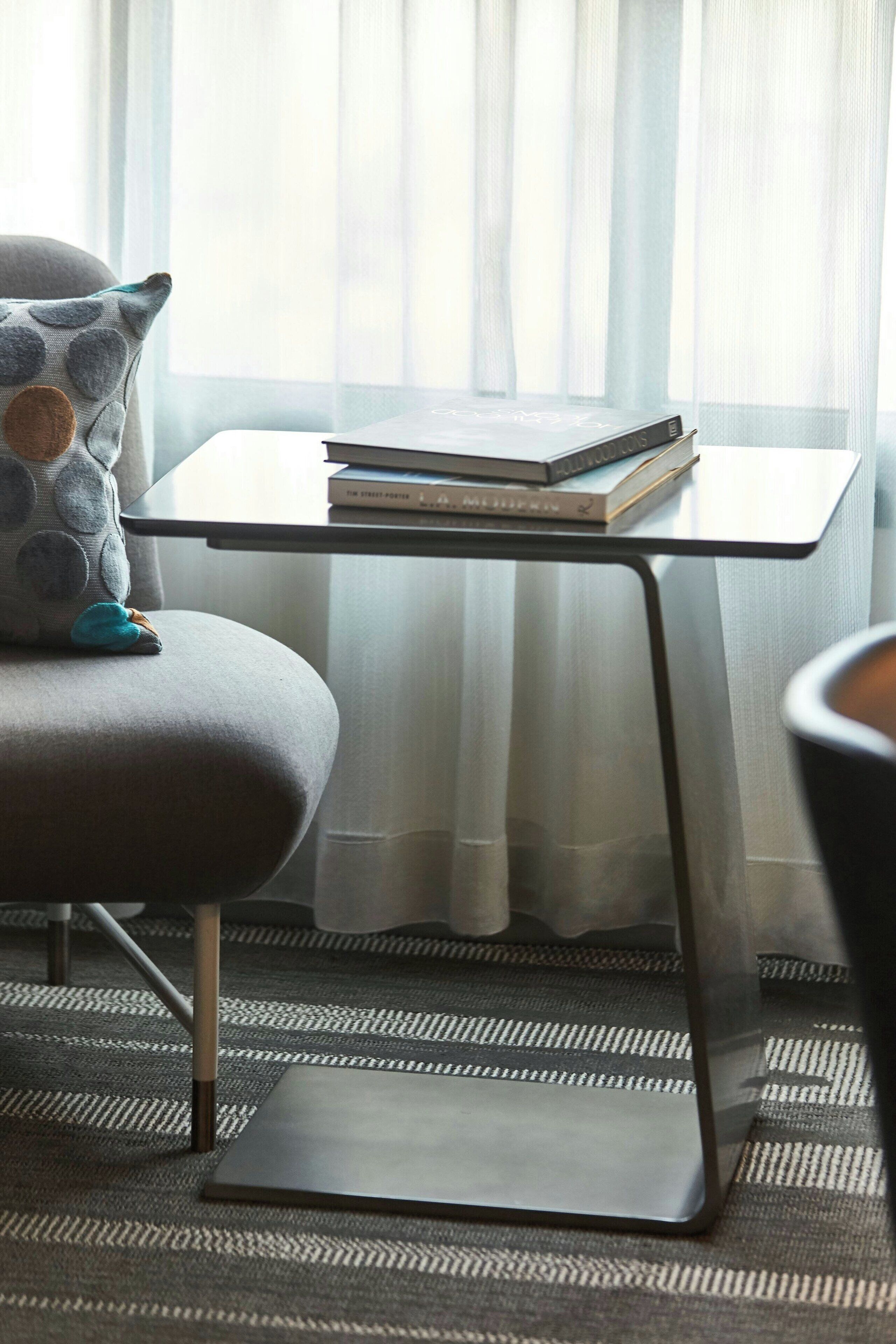 A modern room with a grey armchair, a small table with a stack of books, and a view through sheer curtains.