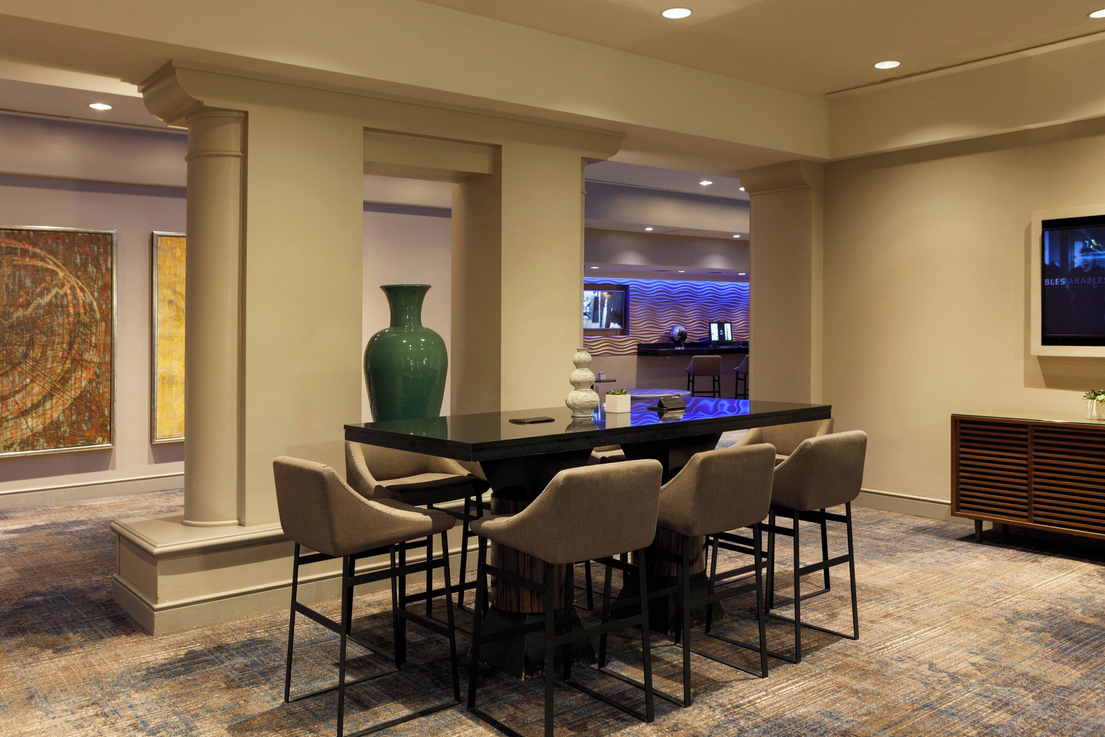 A modern hotel lobby with a black bar table, six bar stools, a large green vase, and artwork on the walls.