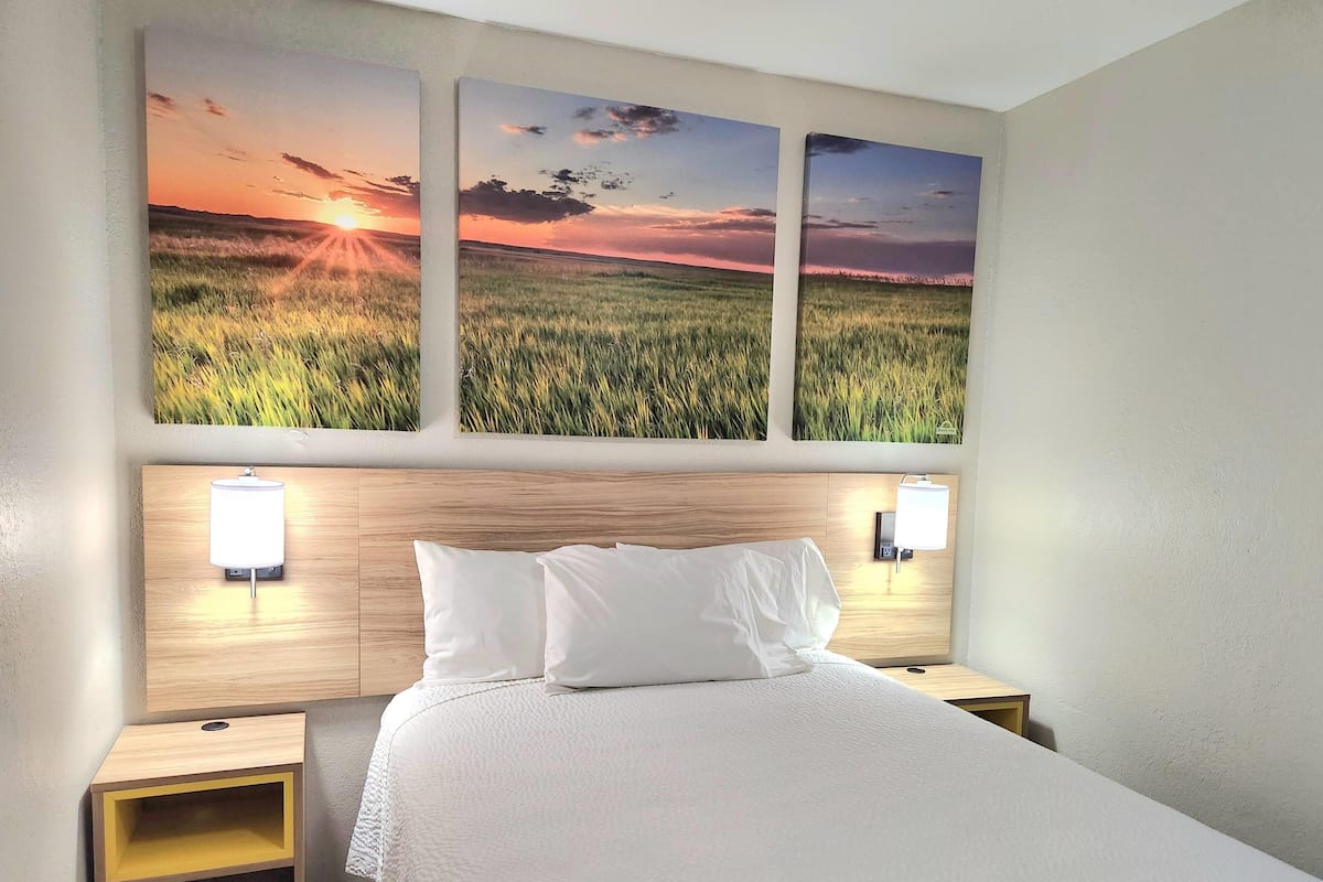 A hotel room with a large bed, two bedside tables, a wooden headboard, and three wall-mounted paintings of a sunset over a field.