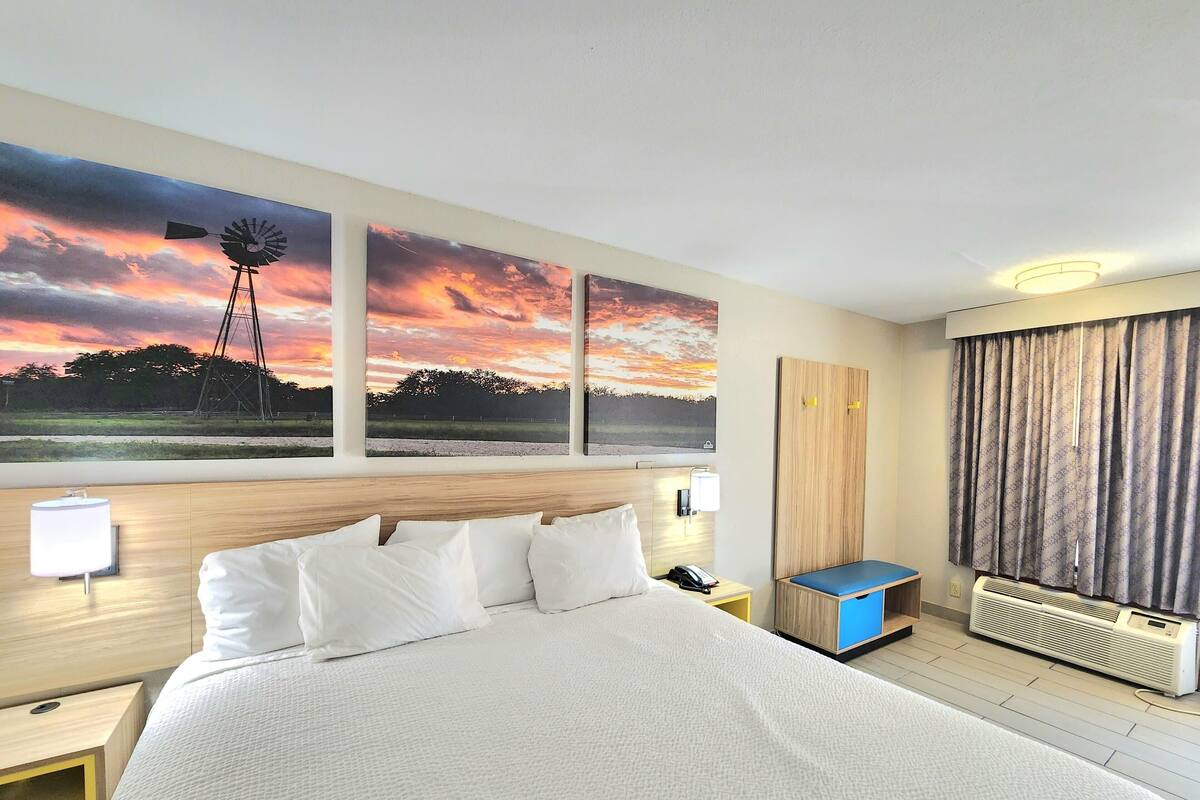 A hotel room with a large bed, a nightstand, a lamp, a desk with a chair, a window with curtains, and a wall-mounted air conditioner.
